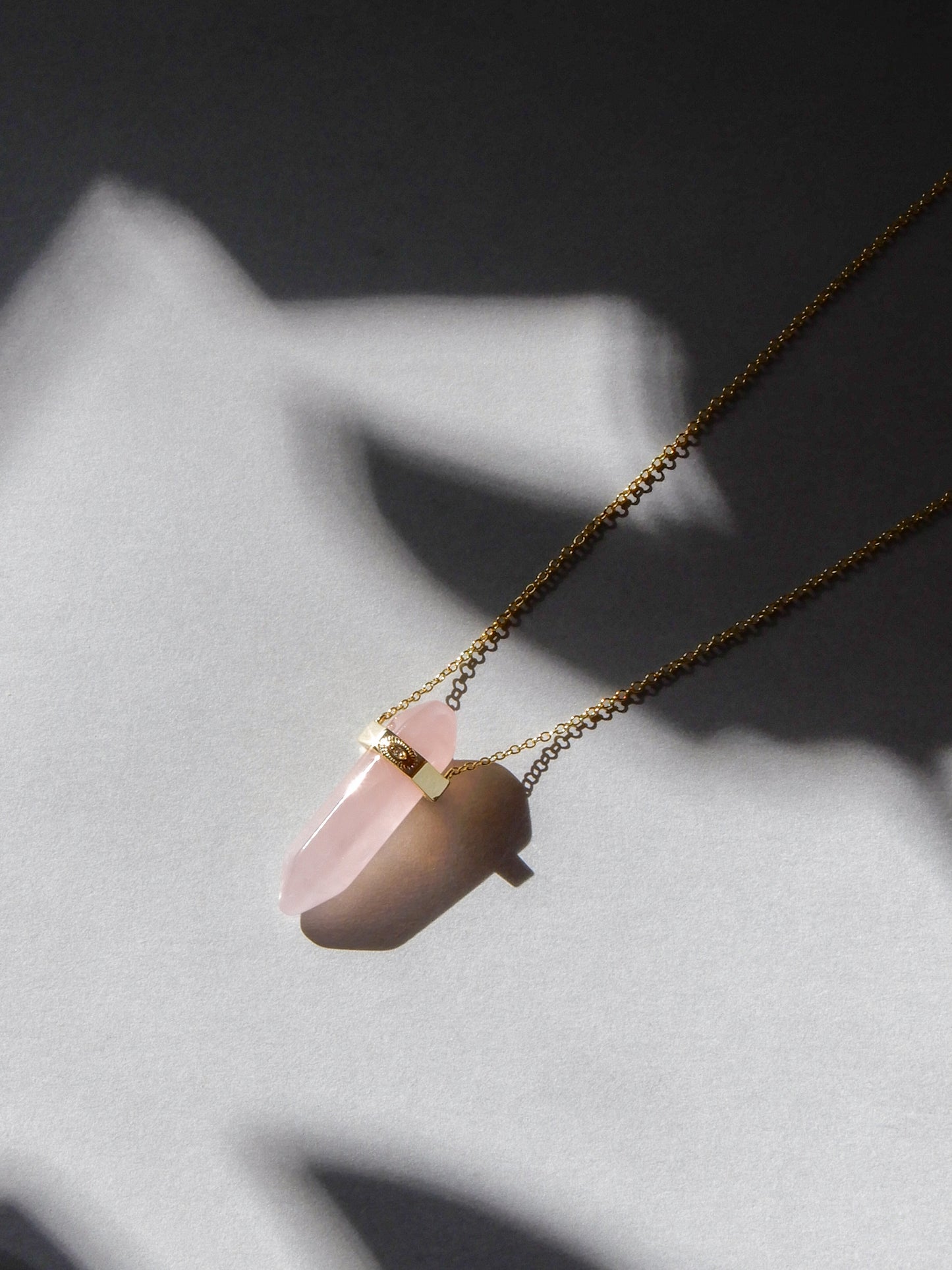 rose quartz crystal with a gold bar wrapper around it. The bar has an evil eye engraved on it. The bar connects to a fine gold chain.