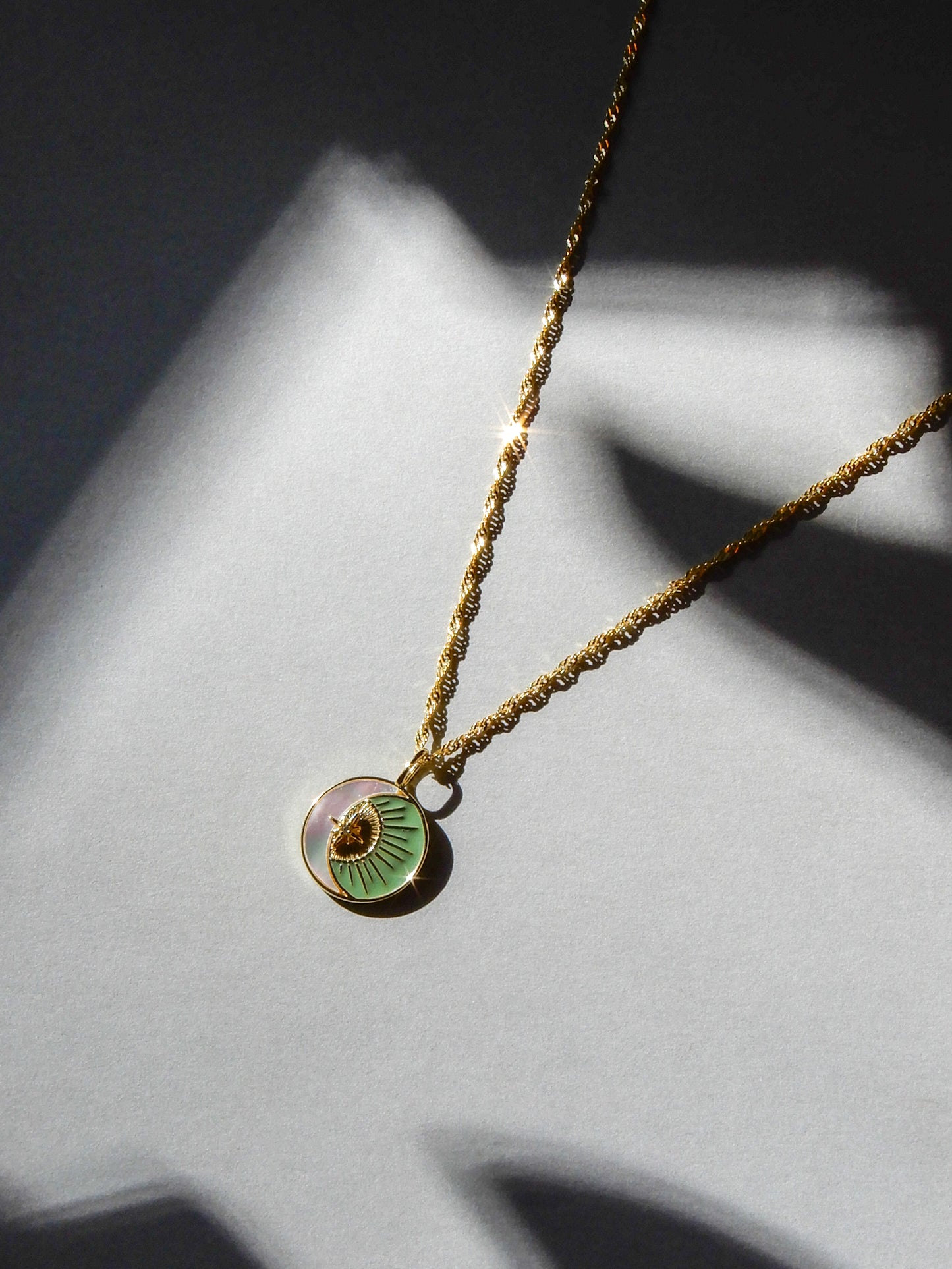 Gold necklace with a green pendant on a twisted chain. The pendant has a pearlescent crescent moon, with a golden sun in the hollow. The necklace is shining in the light, and laid on a gray background.