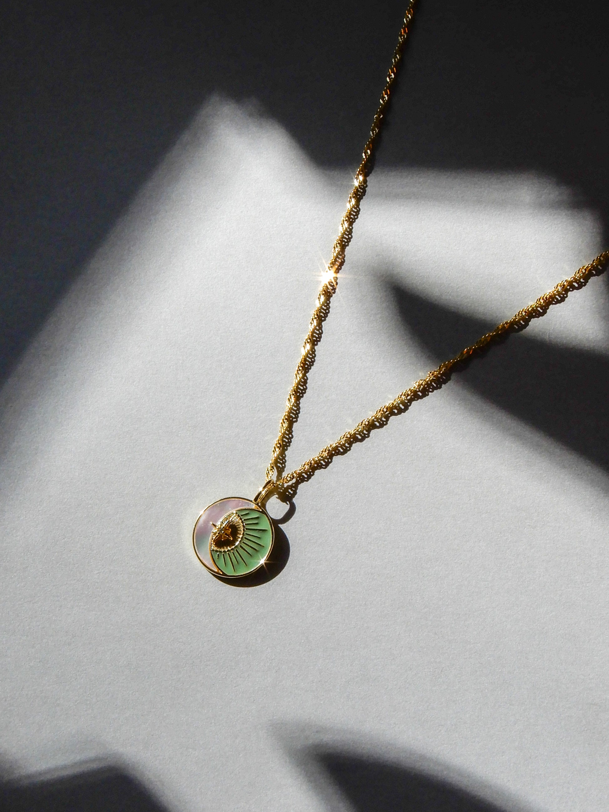 Gold necklace with a green pendant on a twisted chain. The pendant has a pearlescent crescent moon, with a golden sun in the hollow. The necklace is shining in the light, and laid on a gray background.
