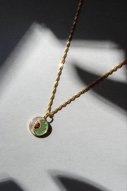 Gold necklace with a green pendant on a twisted chain. The pendant has a pearlescent crescent moon, with a golden sun in the hollow. The necklace is shining in the light, and laid on a gray background.