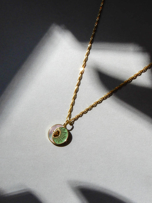 Gold necklace with a green pendant on a twisted chain. The pendant has a pearlescent crescent moon, with a golden sun in the hollow. The necklace is shining in the light, and laid on a gray background.