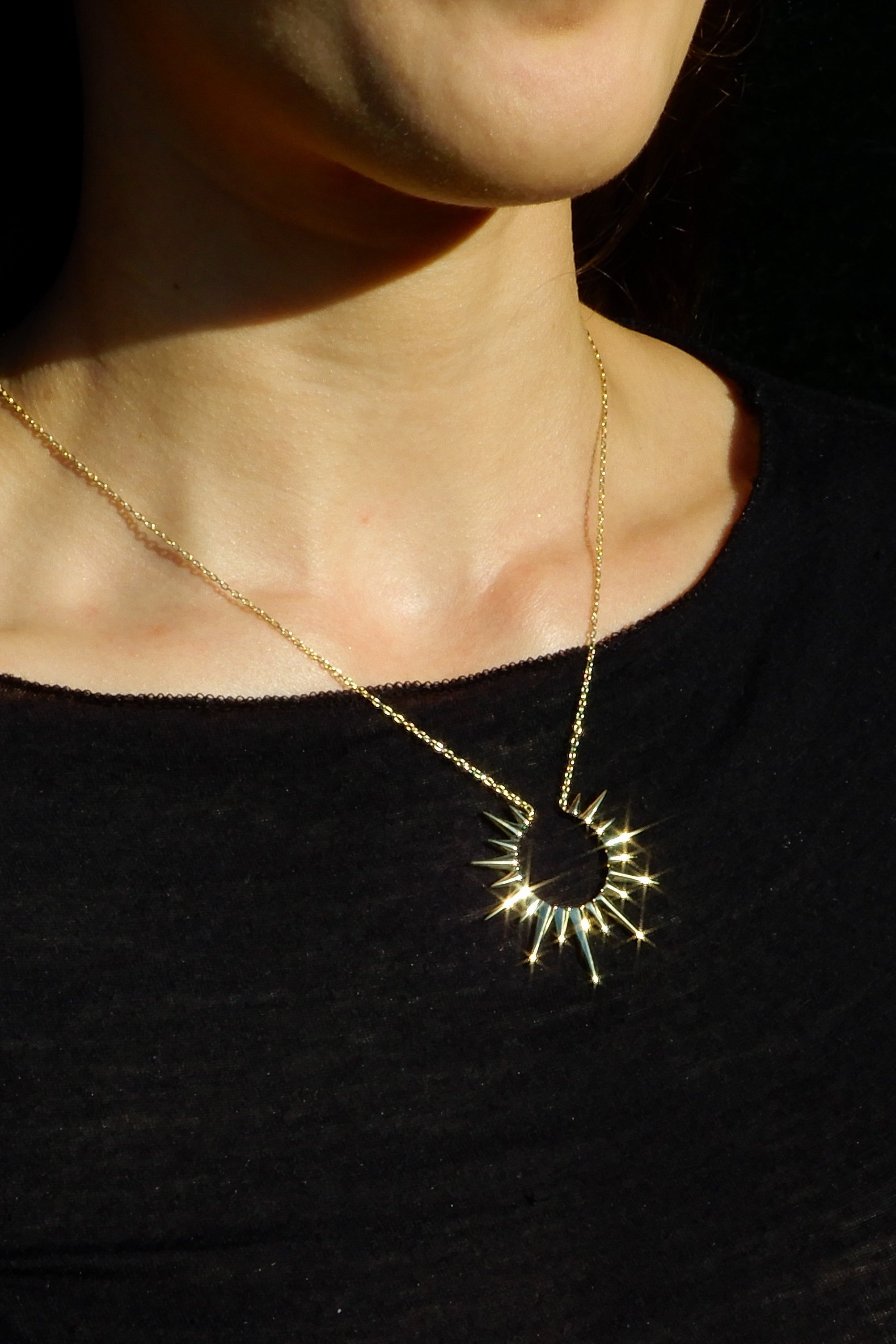 Person wearing a gold necklace with a sunburst pendant against a dark background
