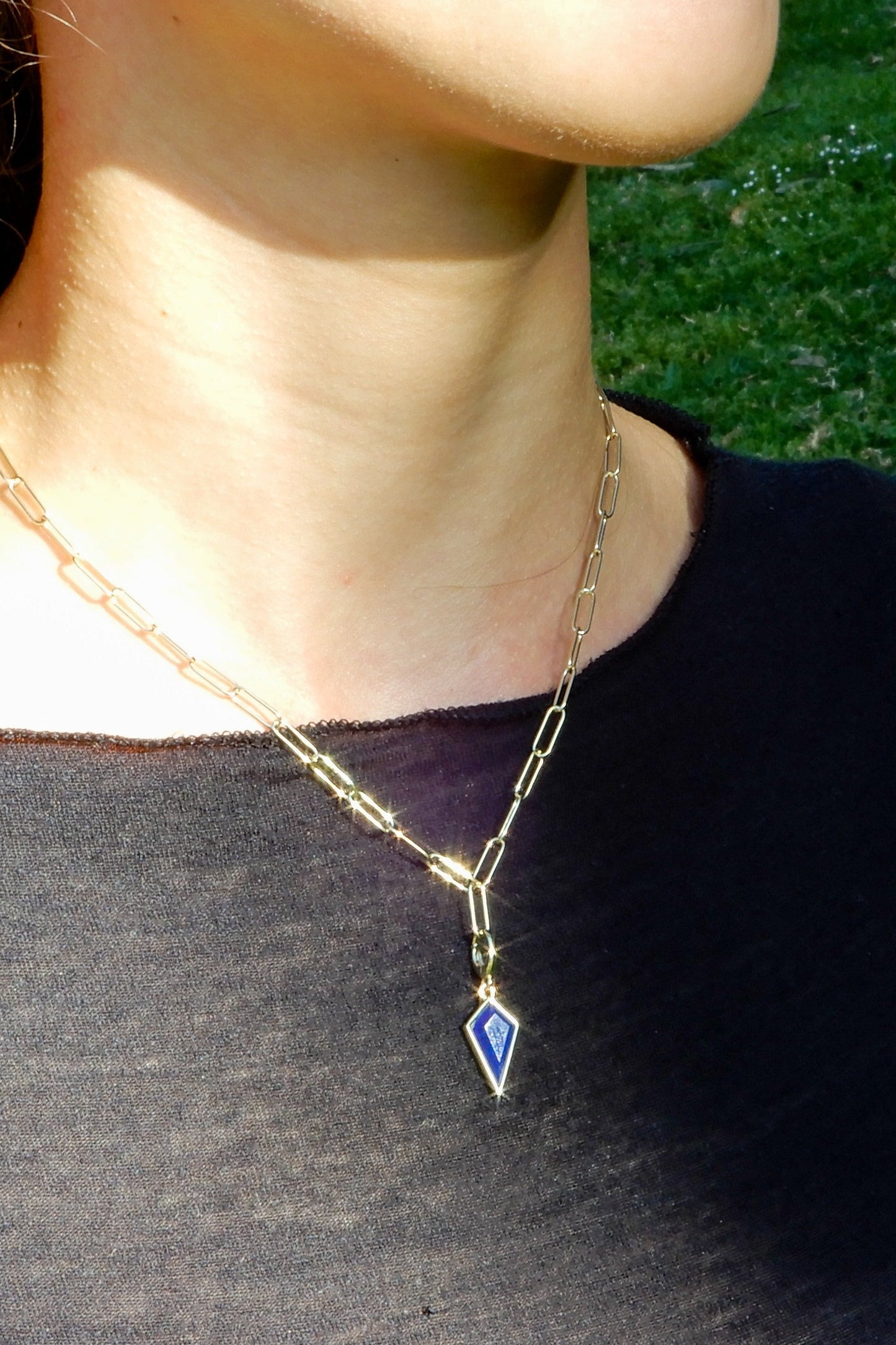 Person wearing a gold necklace with a blue pendant against a grassy background